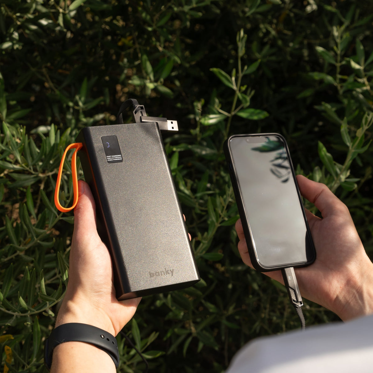Banky powerbank 30.000 mAh met ingebouwde kabels in de ene hand en een telefoon in de andere, buiten tussen het groen