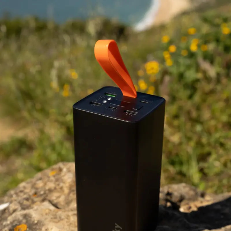 Banky powerbank 50.000 mAh met oranje lus op een rots, met bloemen en zee op de achtergrond