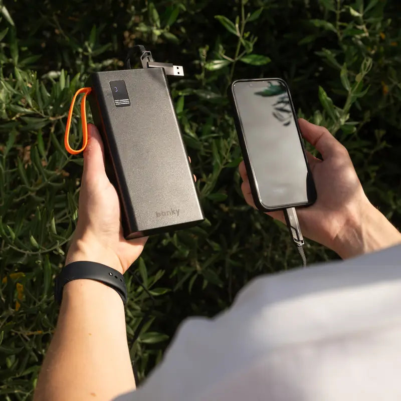 Banky powerbank met ingebouwde kabel laadt een telefoon op, in de hand tussen groene planten
